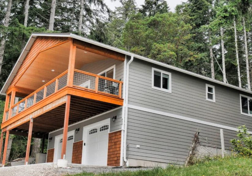 This modern ADU maximizes living space on a sloped lot. The two-story structure features contemporary gray horizontal siding with warm cedar wood accents that create visual interest and contrast. The ground level houses a spacious three-car garage with white doors featuring decorative window panels, while the upper level contains the main living quarters. A dramatic cantilevered balcony extends from the front of the ADU, supported by natural wood posts and eaturing cable railings with a wood frame. The balcony is sheltered by an extended roof line with exposed wood ceiling details, creating an inviting outdoor living space. Multiple windows across both levels ensure abundant natural light throughout the interior. This design represents an excellent example of how ADUs can efficiently use challenging topography while creating attractive, functional additional housing that complements the natural environment.